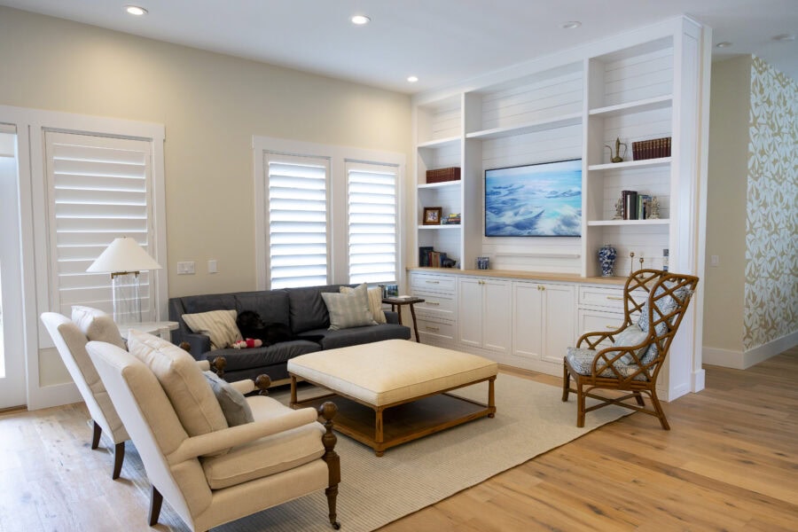Living room with beige walls, white window shutters, a black sofa, beige armchair, wooden coffee table, and built-in white shelves with a TV.