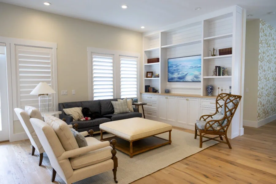 Living room with beige walls, white window shutters, a black sofa, beige armchair, wooden coffee table, and built-in white shelves with a TV.