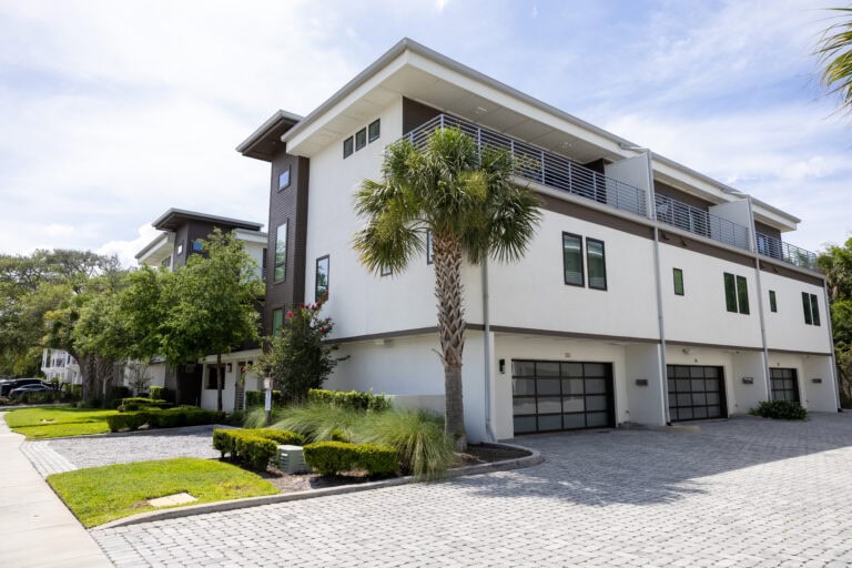 Modern multi-story residential building with white exterior, large windows, and balconies, surrounded by palm trees and landscaped greenery under a partly cloudy sky.