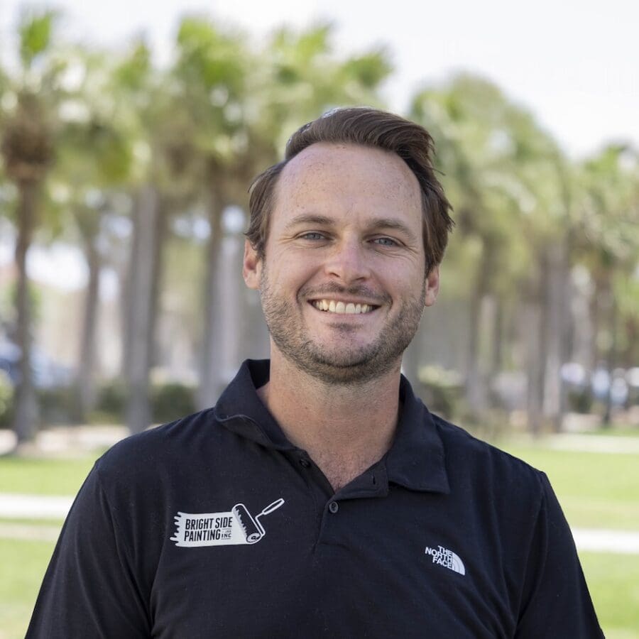 photo of man smiling while standing outdoors wearing a black polo with palm trees in the background