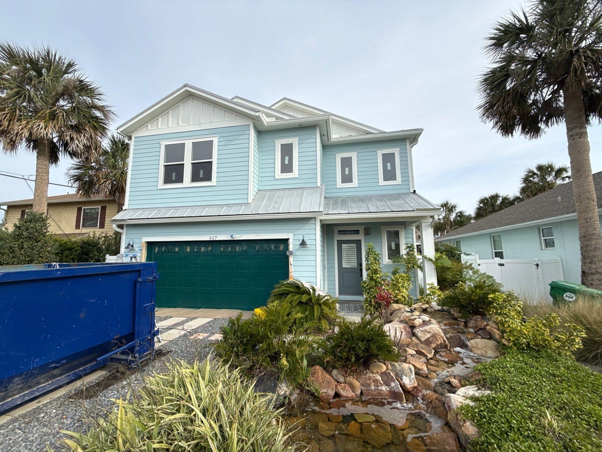 Light blue two-story house with a green garage door, surrounded by palm trees and landscaped garden with rocks and plants.