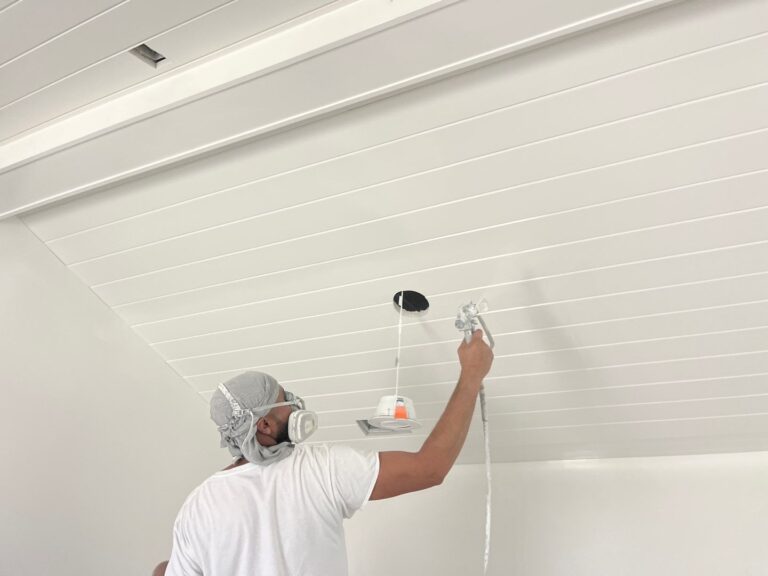 Person wearing a mask and hair cover painting a ceiling with a roller brush.